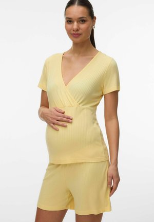 Rase naine kollase soonikkoes lühikeste varrukatega t-särgis ja lühikestes pükstes, õrnalt kõhtu hoidmas, seisab valge tausta ees.