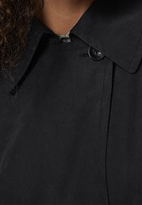 Černý trenčkot s špičatým límcem, velkým zapínáním na knoflíky a hladkou, texturovanou látkou. Má čisté švy podél švů.