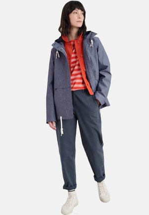 Chaqueta estructurada azul con capucha, camisa de botones roja y pantalones grises. Zapatillas blancas completan el atuendo. Texturas suaves y diseño informal.