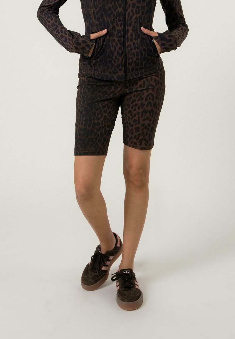 Veste zippée à imprimé léopard brun et short de vélo assorti, fabriqués dans un matériau extensible, associés à des baskets sombres avec des rayures roses.