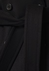 Czarny wełniany płaszcz z fakturowanym materiałem, z przednim zapięciem na guziki i dopasowanym paskiem z tego samego materiału. Szczegóły obejmują ostre kołnierze i dopasowany krój.