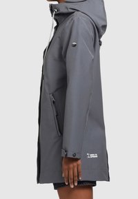 Boční pohled na osobu v šedé voděodolné bundě s kapucí, se zipovými kapsami a knoflíkovými manžetami.