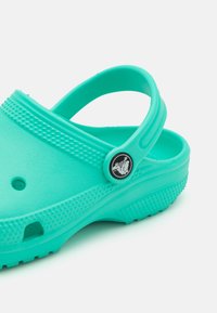 Tirkizne Crocs sa remenom na stražnjoj strani, izrađene od laganog materijala. Imaju odvodne rupe i teksturiranu potplat za prianjanje i udobnost.