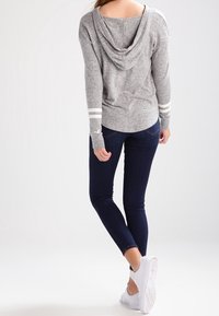 Sudadera gris con capucha, tres rayas blancas en las mangas, combinada con jeans ajustados oscuros y zapatillas blancas, vista desde la parte posterior.