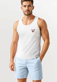 White cotton tank top featuring a small orange and blue logo. Worn with light blue shorts with a smooth texture and side pockets.