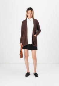 Brown ribbed cardigan with two front pockets, worn over a white button-up shirt, paired with a black skirt and black shoes.