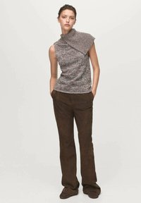 Femme debout avec les mains dans les poches, portant un haut tricoté sans manches gris avec un col asymétrique et un pantalon large marron foncé.