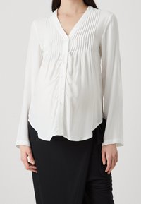 Vit blus med långa ärmar och vertikala veck vid halslinningen, knappstängning och en avslappnad passform, som bärs tillsammans med en svart kjol. Mjuk tygkänsla.