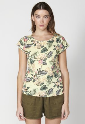 Blusenshirt mit floralem Muster und kurzen Ärmeln, runder Halsauschnitt, aus leichtem gelbem Stoff, mit rosa und grünen Blattmustern. Kombiniert mit olivgrünen Shorts.