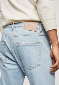 Jeans de mezclilla azul claro con una textura suave, que presentan una etiqueta de cuero en la cinturilla y dos bolsillos traseros con detalles de costura en tono beige.