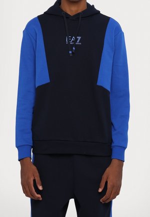 Homme portant un sweat à capuche noir et bleu avec le logo EA7 Emporio Armani sur la poitrine, associé à un pantalon noir avec des bandes latérales bleues.