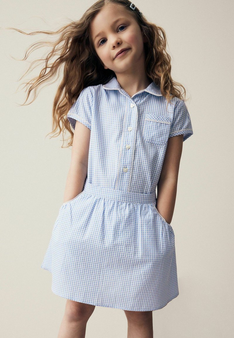 Robe en vichy bleu avec des manches courtes, un devant à boutons et une taille élastique. Elle comprend des poches latérales et une petite poche sur la poitrine.