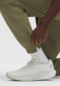 Weißer Sportschuh mit strukturiertem Obermaterial und grauem Logo, kombiniert mit olivgrünen Hosen mit Reißverschluss am Saum, getragen über einer weißen Socke.