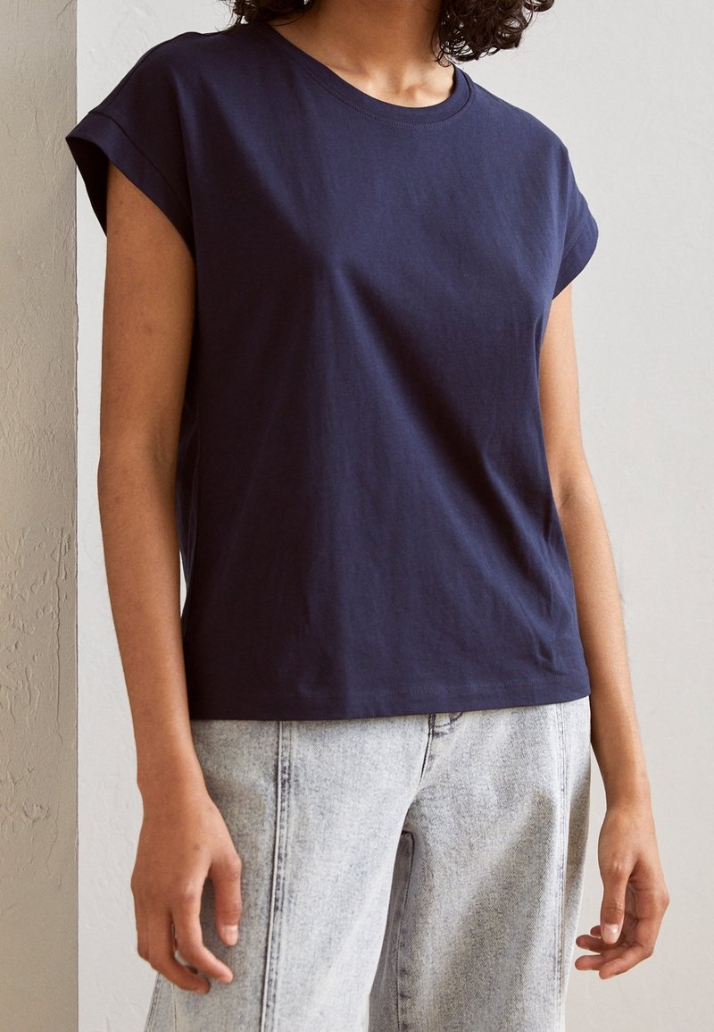 Personne portant un t-shirt ample bleu foncé à manches courtes et un jean taille haute gris clair, debout contre un mur uni.