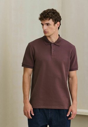 Ung mand med krøllet hår iført en brun poloshirt med korte ærmer og mørke bukser, kigger mod sin venstre side mod en ensfarvet baggrund.