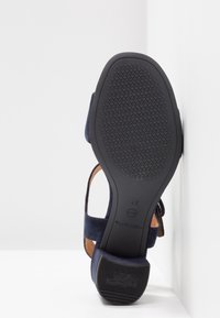 Sandale en suède bleu marine avec une semelle en caoutchouc noir, présentant un motif texturé et un design de brides. Étiquette de taille visible sur la semelle intérieure.