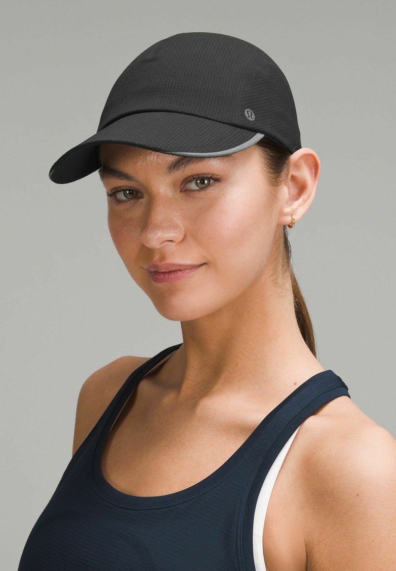 Gorra atlética negra con una superficie texturizada, visera curva y un acento reflectante. Combinada con una camiseta sin mangas oscura. Diseño limpio y moderno.