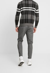 Pull gris à carreaux avec des rayures noires et blanches sur les épaules, associé à un pantalon de jogging gris et des baskets blanches. Poches latérales visibles.