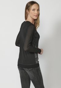 Haut noir à manches longues avec des manches en maille texturée, présentant un motif floral sur le côté, terminé par un ourlet et des poignets côtelés.
