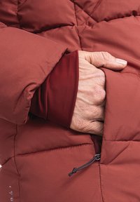 Rote gepolsterte Jacke mit gestepptem Design. Eine Hand ist in der seitlichen Tasche platziert. Der Innenärmel verfügt über eine passgenaue rote Schicht.