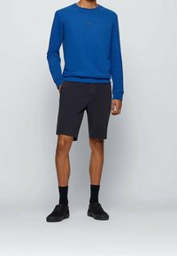 Blå sweatshirt med rund hals och logo, i kombination med mörkgrå shorts. Modellen bär svarta strumpor och svarta skor. Enkelt design, avslappnad passform.