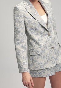 Femme portant un blazer texturé gris clair à motifs avec deux boutons et un short assorti, avec un top blanc visible en dessous.