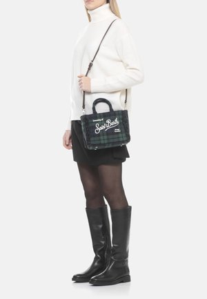 Donna con dolcevita bianco, gonna nera, calze trasparenti e stivali neri al ginocchio, che tiene una borsa Saint Barth a quadri verdi con tracolla.