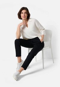 Cremefarbener V-Ausschnitt-Pullover aus strukturiertem Stoff, kombiniert mit schmalen schwarzen Hosen und weißen Sneakers, sitzt auf einem weißen Stuhl.