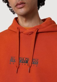 Detailní záběr osoby v oranžové mikině s tkaničkami a černým logem "Napapijri Geographic" na hrudi.