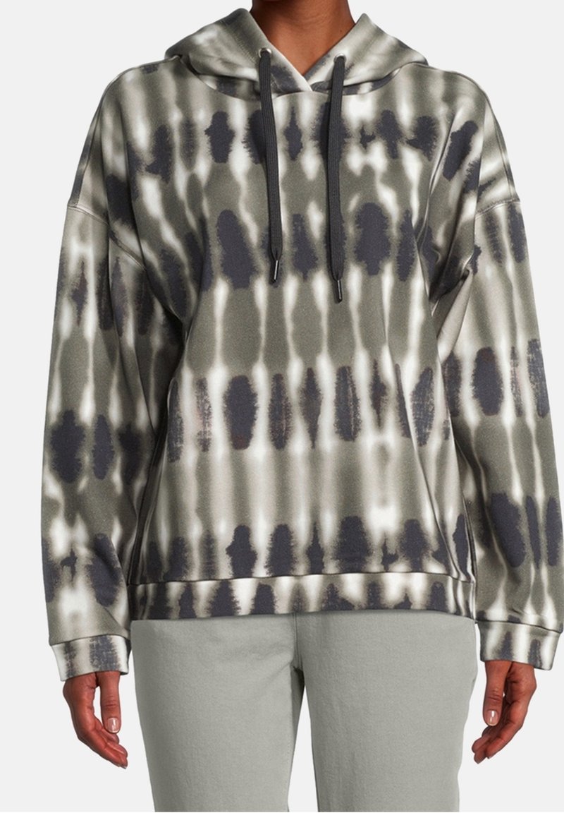 Personne portant un sweat à capuche tie-dye ample dans des tons de gris et blanc avec des cordons noirs, associé à un pantalon gris clair.