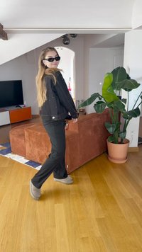 Jaqueta de denim preta, calças pretas ajustadas e botas bege. Cabelo castanho longo e óculos de sol escuros. Ambiente interior com um sofá castanho texturizado.