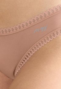 Nærbillede af beige undertøj med blonder på huden, med "sloggi" logo på stoffets side.