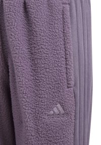 Lila fleece-joggers med strukturerad tyg, med en Adidas-logotyp på benet. Vertikala ränder syns på sidan.