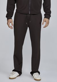 Brown athletic pants made of smooth fabric, featuring a drawstring waist and side pockets. Paired with white sneakers.