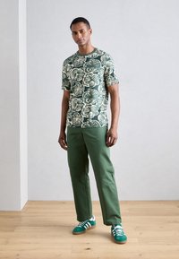 Green floral-patterned short-sleeve shirt, paired with green straight-leg pants and green athletic shoes with white stripes. Wooden floor background.
