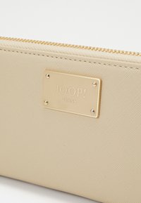 Portafoglio beige testurizzato con hardware dorato, caratterizzato da una piastra metallica rettangolare incisa con "JOOP! JEANS" fissata da quattro borchie.