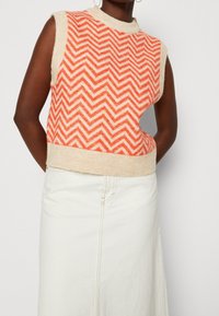 Ærmeløs strikket sweater med et markant chevron-mønster i orange og creme, med en ribbet creme halskant og matchende bundtrim.