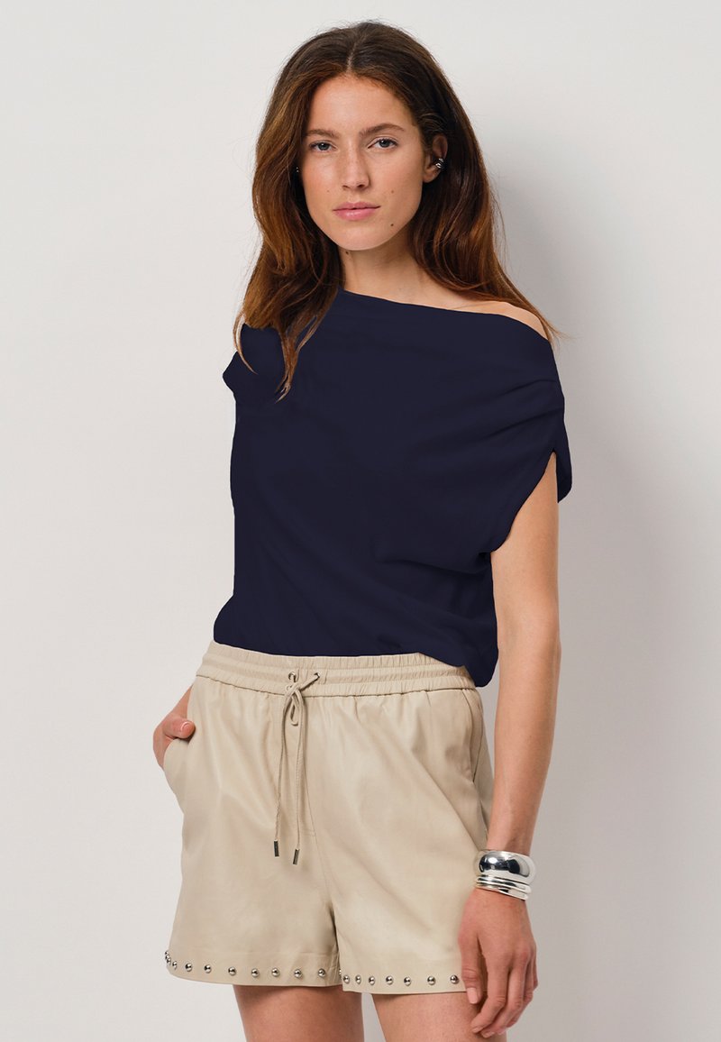 Marine off-shoulder top gecombineerd met beige shorts met een trekkoord en studs aan de zoom. Model met lang haar.