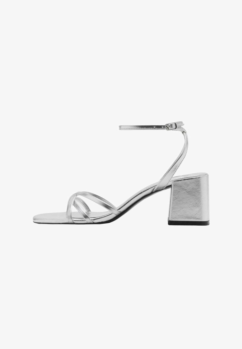 Zilveren sandaal met blokhak, voorzien van dunne kruislingse banden en een verstelbare enkelband. Gladde metallic afwerking.