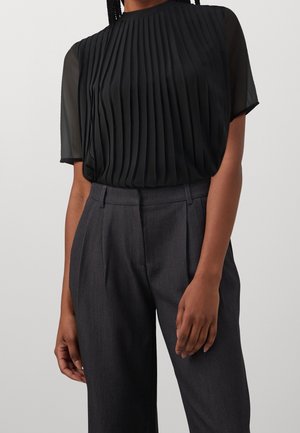 Femme portant un chemisier noir plissé à manches courtes avec des manches transparentes et un pantalon tailleur gris foncé taille haute.