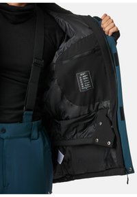 Gepolsterte Jacke mit schwarzem Innenfutter und mehreren Taschen. Verfügt über ein STX-Tag, Reißverschluss und Klettverschlüsse. Außen in Blaugrün mit schwarzen Akzenten.