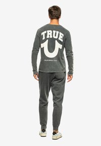 Grijze lange mouwen shirt met "TRUE" en een groot symbool op de achterkant gedrukt. Gecombineerd met bijpassende grijze broek en casual schoenen.