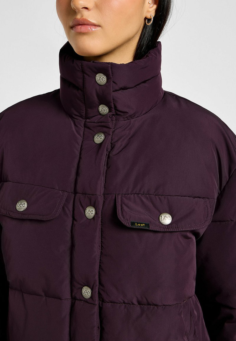 Paarse pufferjack met een hoge kraag, drukknoopsluiting en twee voorzakken. Kenmerkt metalen knopen en een klein logo-label op de zak.