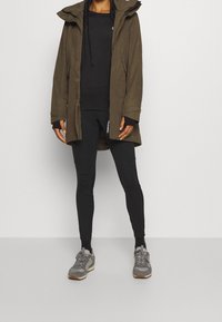 Olivgrön parkas med huva, med dragkedja och knappstängning, i kombination med svart långärmad tröja och leggings, samt grå sneakers.