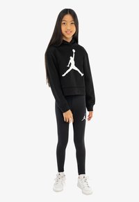 Črna kapuca s belim logotipom Jordan, v kombinaciji s črnimi legicami, ki imajo majhen logotip. Model nosi bele superge s črnimi podrobnostmi.