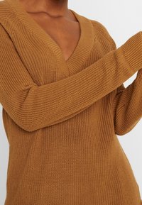 Strickpullover in einer warmen, bernsteinfarbenen Nuance. Mit einem tiefen V-Ausschnitt, langen Ärmeln und strukturiertem Stoff für zusätzliche Details.
