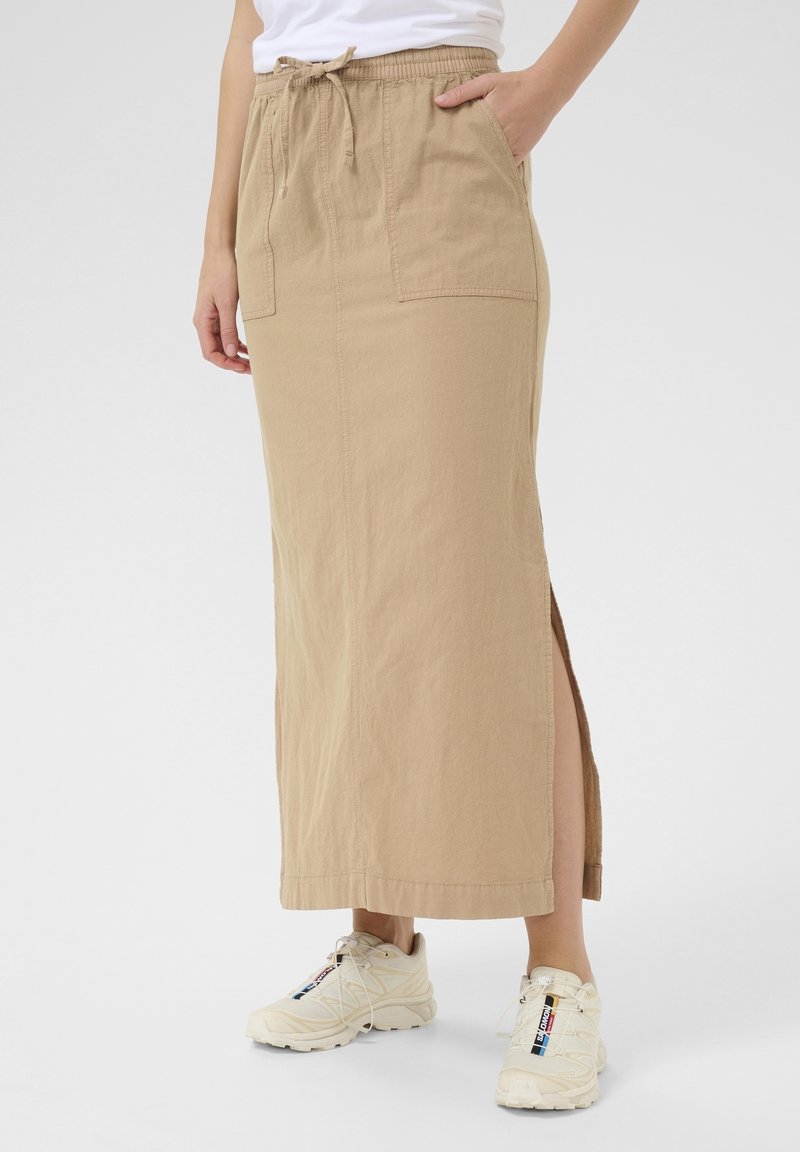 Jupe maxi beige avec une ceinture élastique, un cordon de serrage, des poches latérales et une fente haute. Associée à des chaussures de sport de couleur claire.