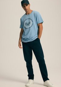 Jasnoniebieska koszulka z krótkim rękawem z grafiką stowarzyszenia tenisowego, połączona z ciemnozielonymi spodniami i białymi trampkami, tworząc swobodny strój.