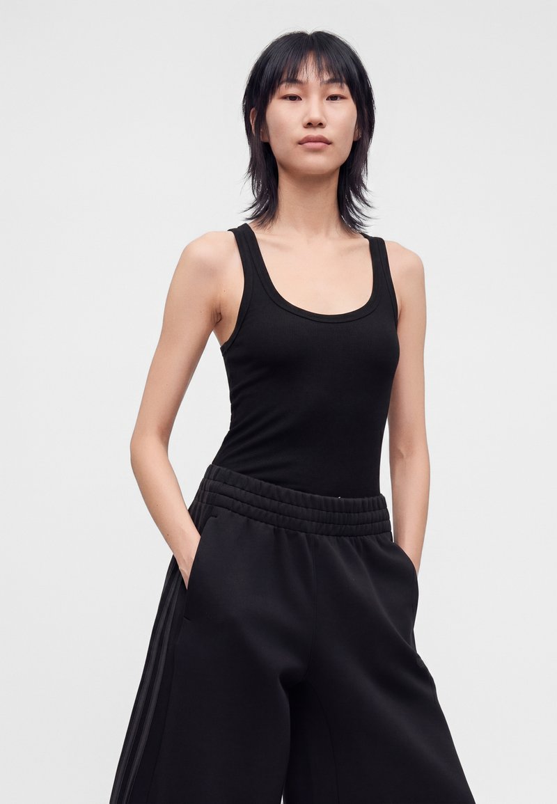 Personne élancée aux cheveux courts noirs portant un débardeur noir sans manches et un pantalon noir ample, les mains dans les poches.
