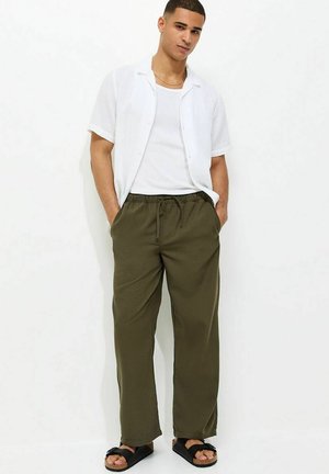 Młody mężczyzna stoi z rękami w kieszeniach, ubrany w białą koszulę z krótkim rękawem, biały podkoszulek, oliwkowe spodnie i czarne sandały.